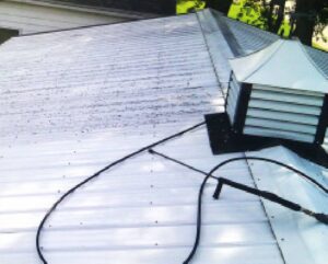 A white metal roof with a vent and a black hose lying across its surface. Trees are visible in the background.