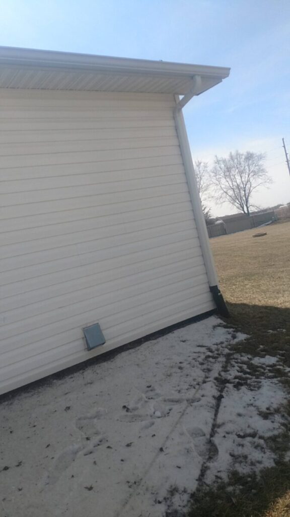 White siding of a house with a vent, concrete area, patchy snow, and grass yard with a tree in the background.