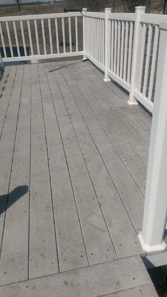A weathered wooden deck with white railings, shown in daylight with a rural landscape in the background.