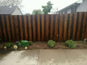 A green toy tractor with a trailer sits near plants by a wooden fence on a wet concrete patio.
