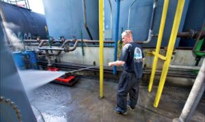 Person using a high-pressure water hose to clean an industrial area with pipes and tanks in the background.