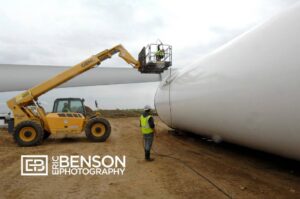 A worker on a lift inspects a wind turbine blade while another stands nearby at a construction site.