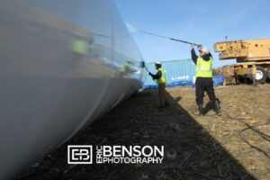Two workers in safety vests clean a large cylindrical structure outdoors with equipment, beside a blue shipping container.