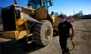 A man uses a pressure washer to clean the front wheel and side of a large yellow construction vehicle outdoors.