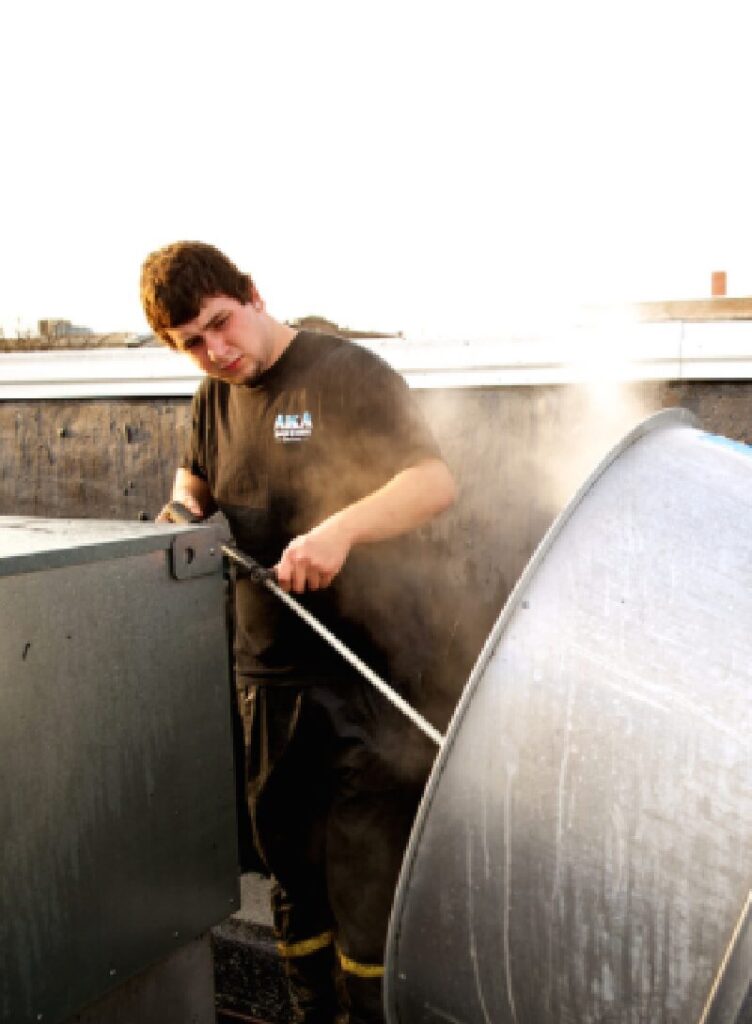 A man in work clothes uses a tool to repair or clean a large industrial fan or vent on a rooftop.