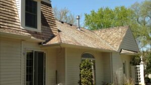 House exterior with tan wood shingle roof, cream siding, several windows, and trees in the background.