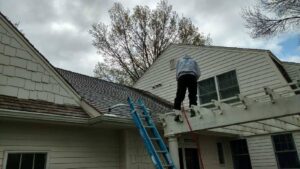 Person standing on a roof near a ladder, working on a two-story white house under cloudy skies.
