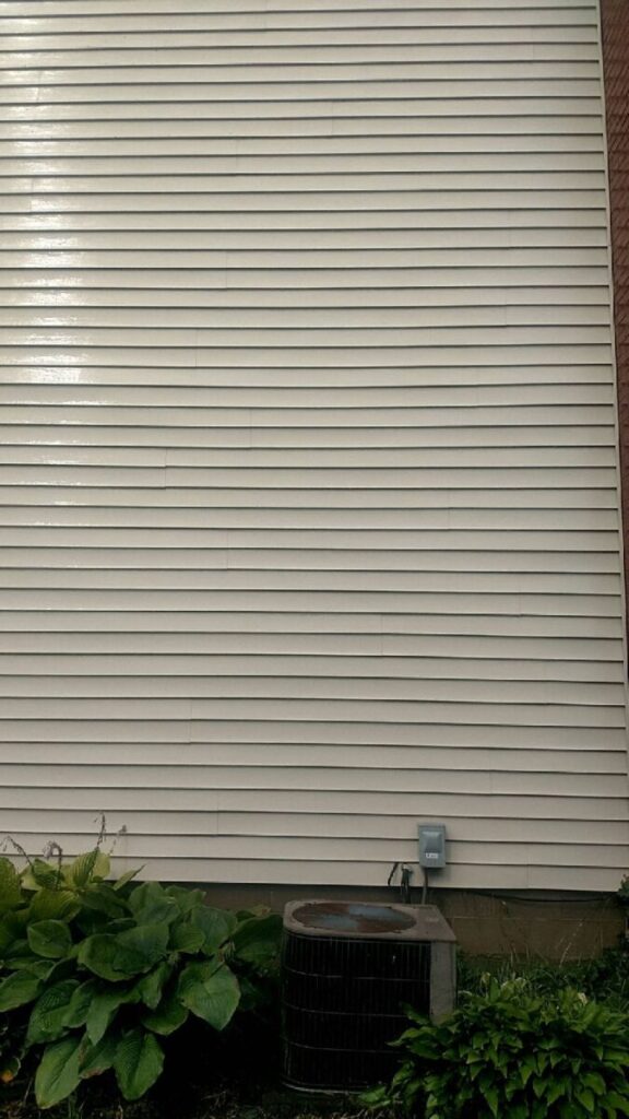Beige vinyl siding wall with an outdoor air conditioning unit and green plants at the base.