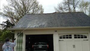 Person pressure washing the roof of a house with an attached garage and trees in the background.