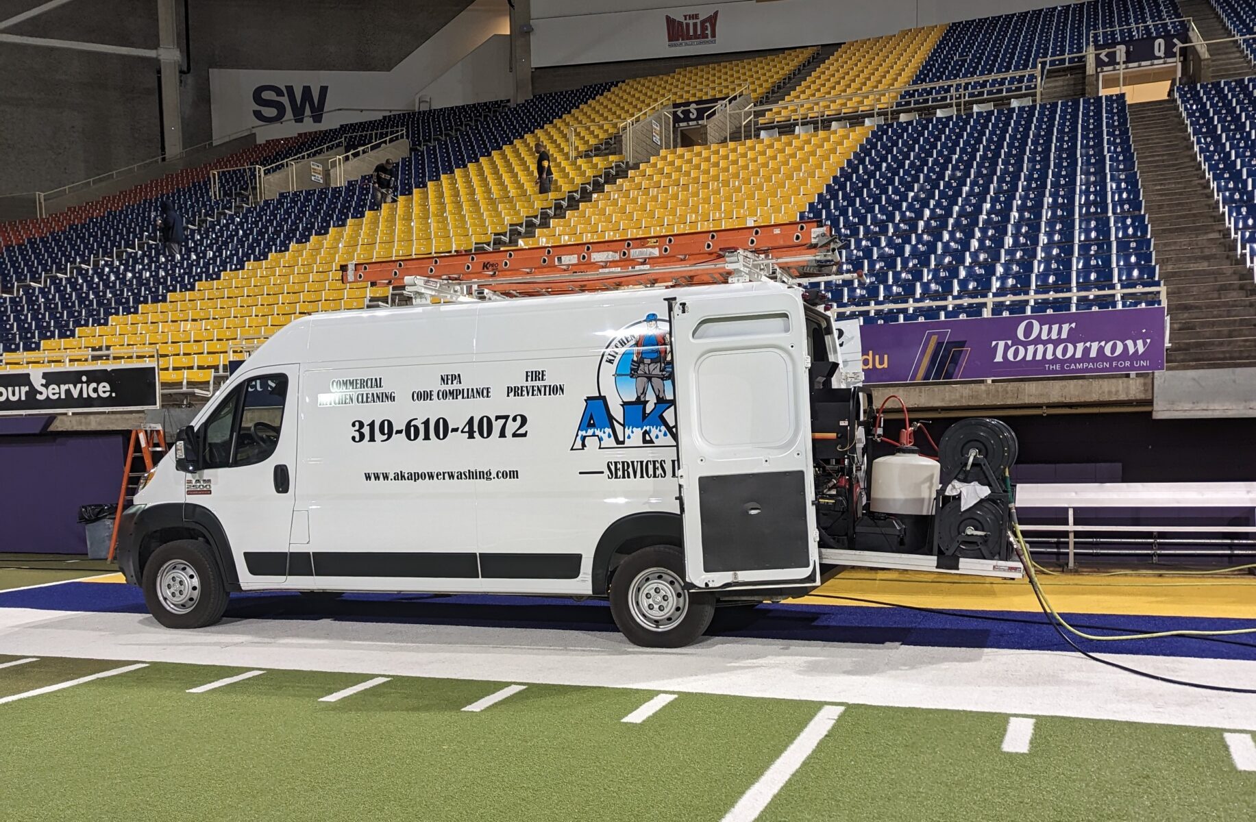 A white service van with cleaning equipment is parked on artificial turf inside a large indoor stadium.